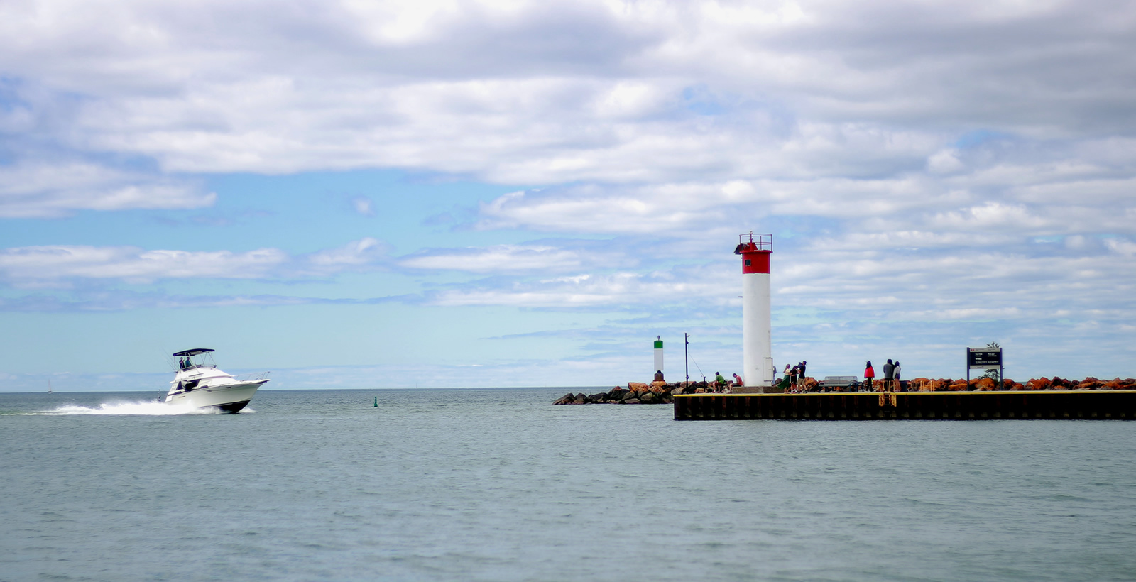 A picture of a boat on Lake Ontario moving towards a pier which has a red lighthouse and people on it on a bright summer day.
